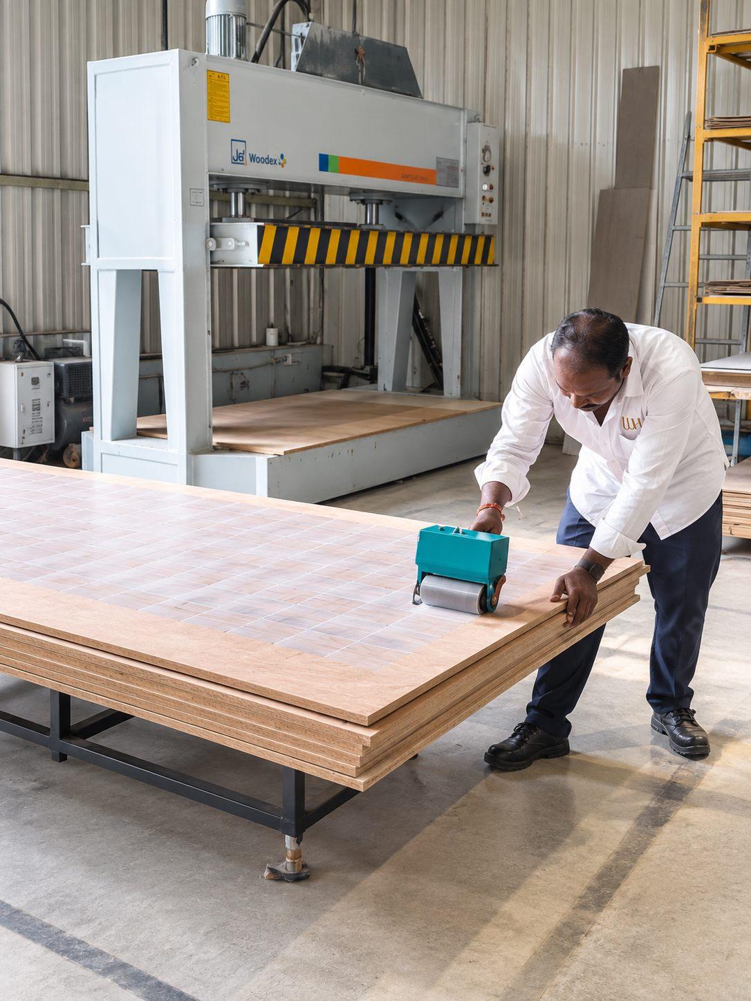 Craftsman rolling an even coat of glue onto a plywood panel before laminate pasting, with the cold press behind