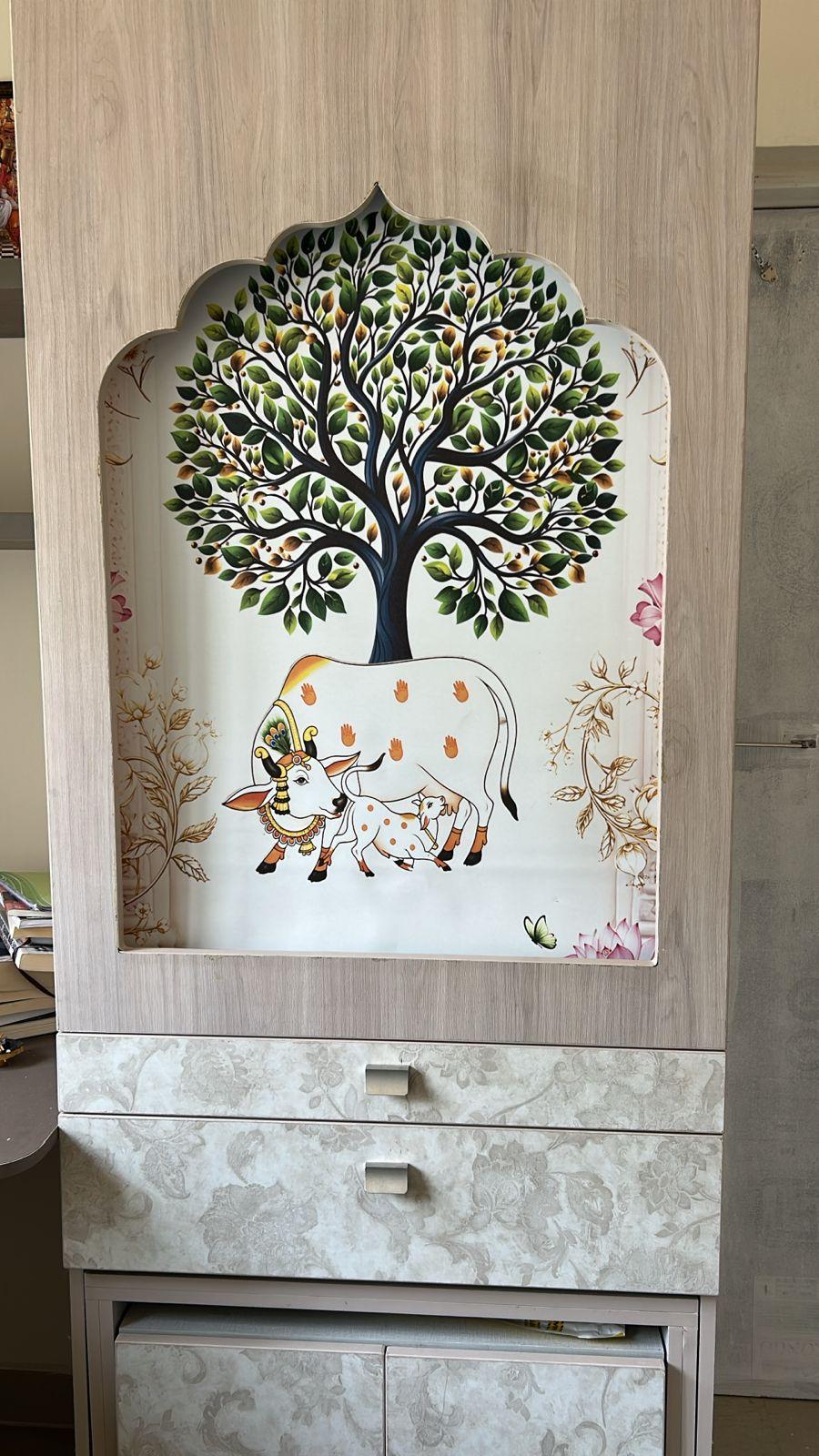 Pooja unit with a Pichwai-style tree and cow artwork in an arched niche, above floral laminate drawers